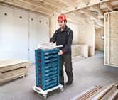 在建的建築物中，配戴安全設備的人員用輪式推車運送堆放的工具箱。
