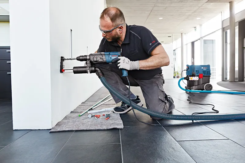 一名配戴安全設備的人員正在用除塵裝置在牆上鑽孔。.