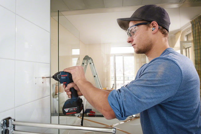 一名穿著安全裝備的工人正在浴室的磁磚牆上鑽孔。.