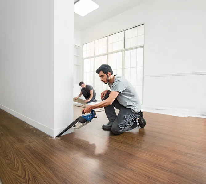 GAS 18V-1 一個人用無線鋸在牆邊切割地板,另一個人則安裝地板鑲板。.