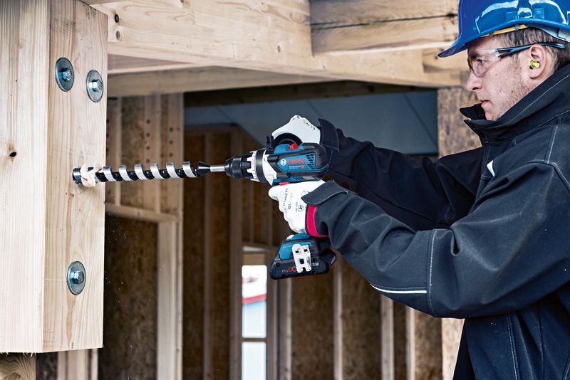 建築工地上，一名配戴安全設備的工人正在向木樑上鑽孔。.