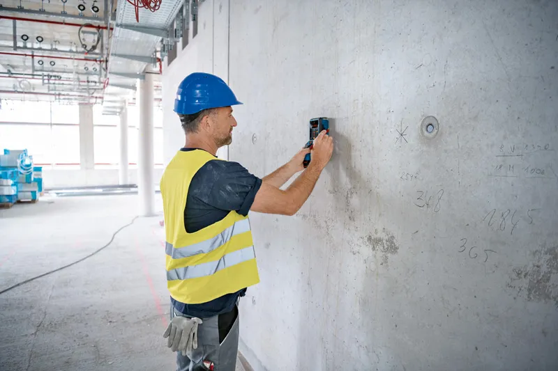 一名配戴安全設備的人員正在使用偵測器掃描混凝土牆。.