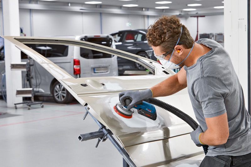 汽車修理廠裡，一名配戴安全設備的人員正在用電動砂光機打磨車門。.