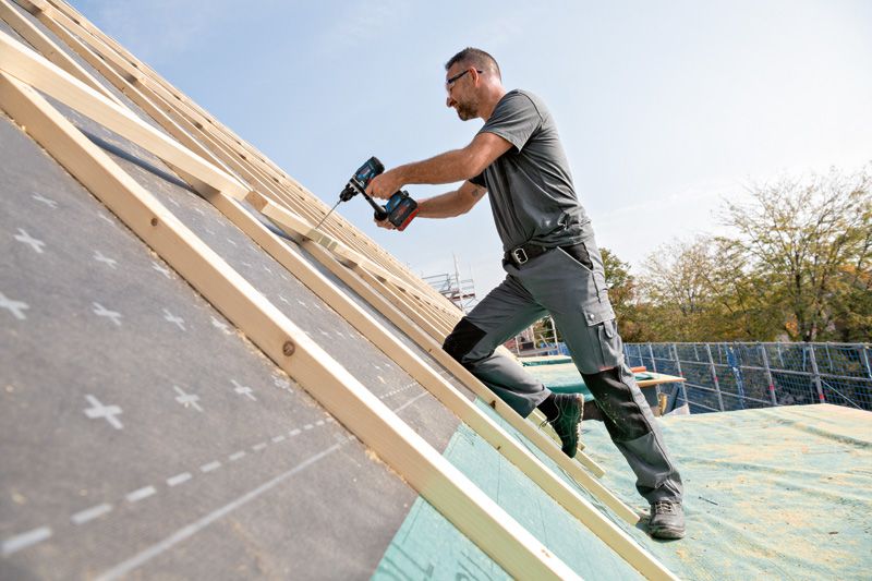 一名佩戴安全設備的人員使用無線電鑽將木頭固定在傾斜的屋頂上。.