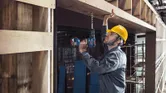 建築工地上，一名配戴安全設備的工人正在向木樑上鑽孔。