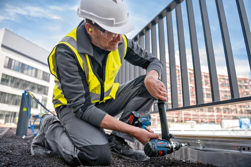一名配戴安全設備的人員使用無線組合鑽在金屬柵欄框架上鑽孔。.