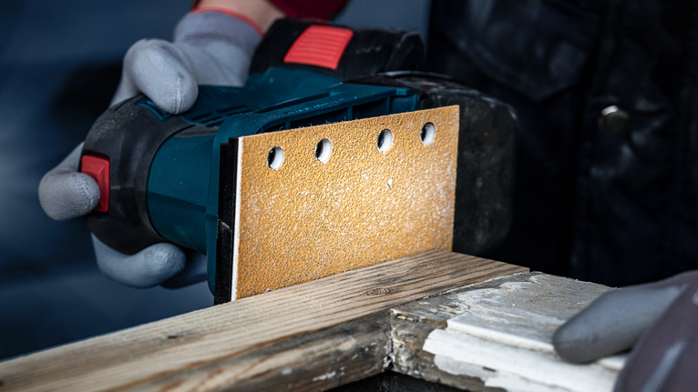 一名配戴安全設備的人員使用手持式電動砂光機打磨木材。