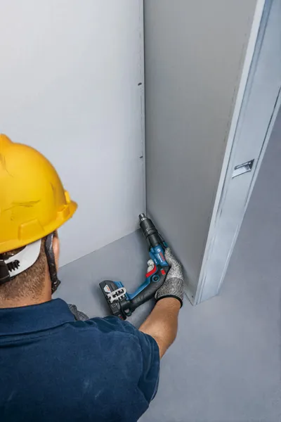 一名配戴安全設備的人員正在用無線螺絲起子固定乾牆。.