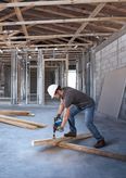 建築工地上，一名配戴安全設備的工人正在向木樑上鑽孔。
