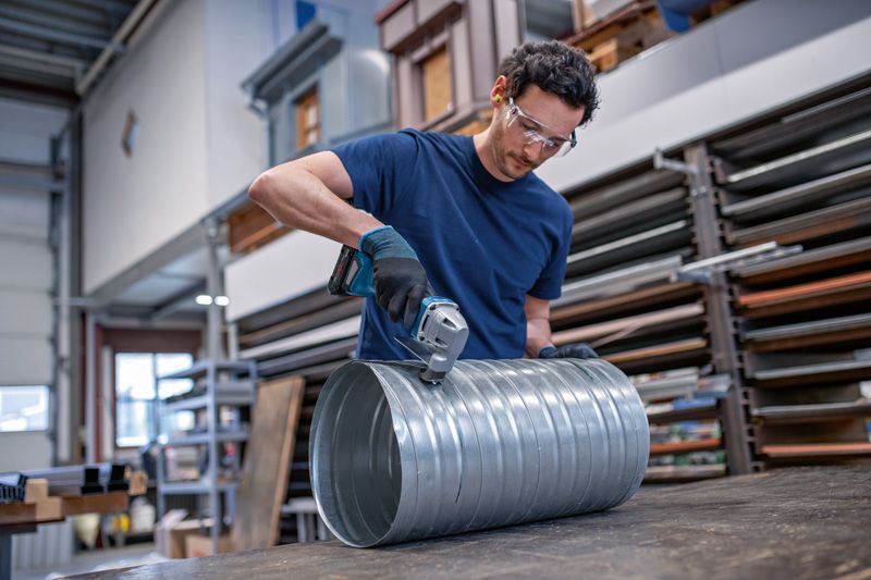 一名配戴安全設備的人員正在使用無線金屬剪剪一條大型金屬管道。.