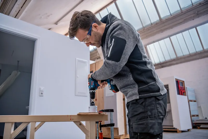 一名配戴安全設備的人使用無線電鑽在木頭上鑽孔。.