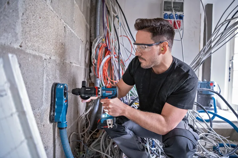 一名配戴安全設備的人員正在用除塵器在混凝土牆上鑽孔。.