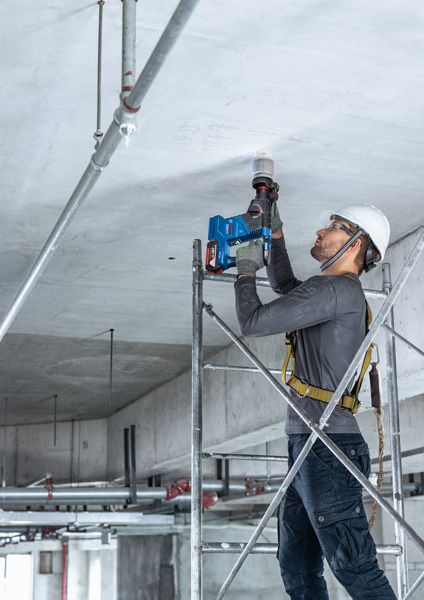 一名配戴安全設備的人員站在鷹架上，向混凝土天花板上鑽孔。.