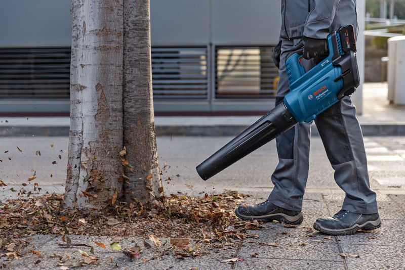 一名穿戴安全裝備的人員使用無線吹葉機清除樹木附近的落葉。
