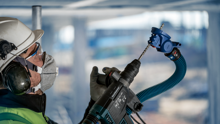 一名佩戴安全設備的人員使用帶有除塵器的電動工具在天花板上鑽孔。