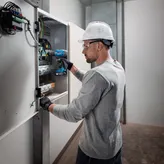 一名佩戴安全設備的人員使用無線電鑽在電氣面板上工作。