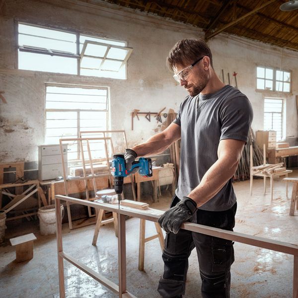 一名配戴安全設備的人員在車間內鑽入木製框架。.