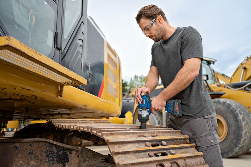 一名配戴安全設備的人員在挖土機履帶上使用無線衝擊扳手。.