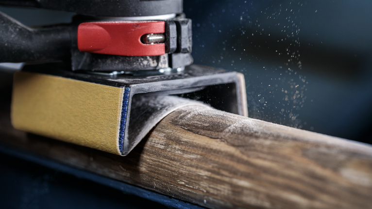 軌道砂光機正在打磨圓形木樑，木屑顆粒清晰可見。