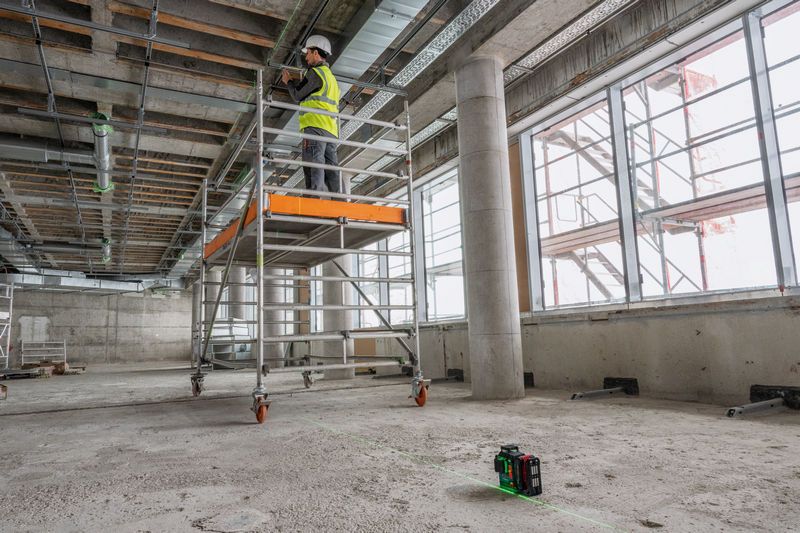 一名佩戴安全設備的人員在鷹架上工作，並使用雷射調平工具投射線條。.