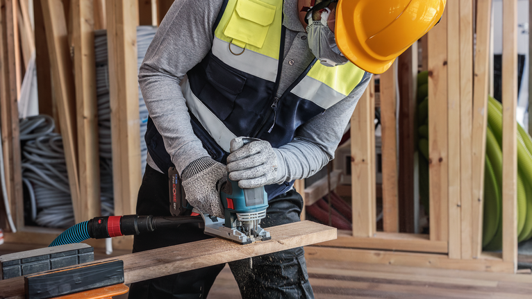 在建築工地上，一名配戴安全設備的人員正在使用電動工具切割木材。