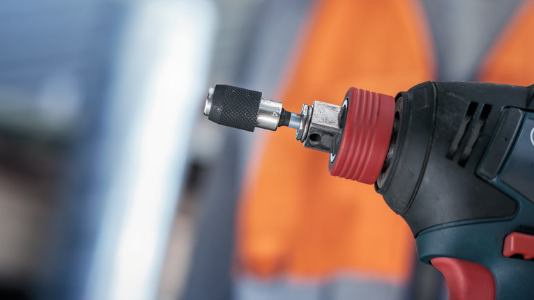 圖中顯示了帶有金屬鑽頭的電動工具，背景中的人們佩戴著安全設備。