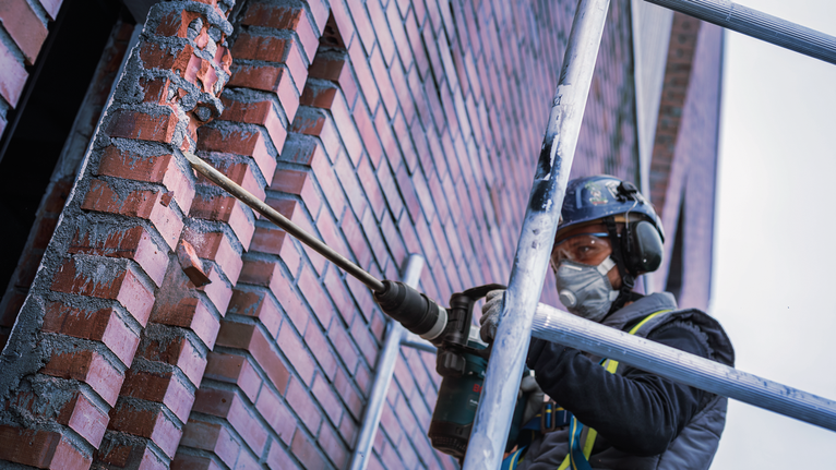 一名身穿安全設備的人員在鷹架上鑽入磚牆。