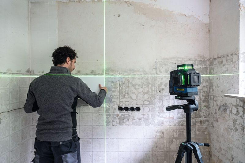 一個人使用三腳架上的綠色雷射調平工具對準牆上的瓷磚。.