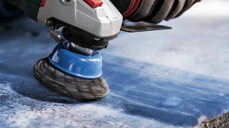 PRO Metal clean 碗形刷,75 x 0.3 mm,螺紋 一名佩戴安全設備的人使用帶有鋼絲杯刷的電動工具在藍色表面上工作。