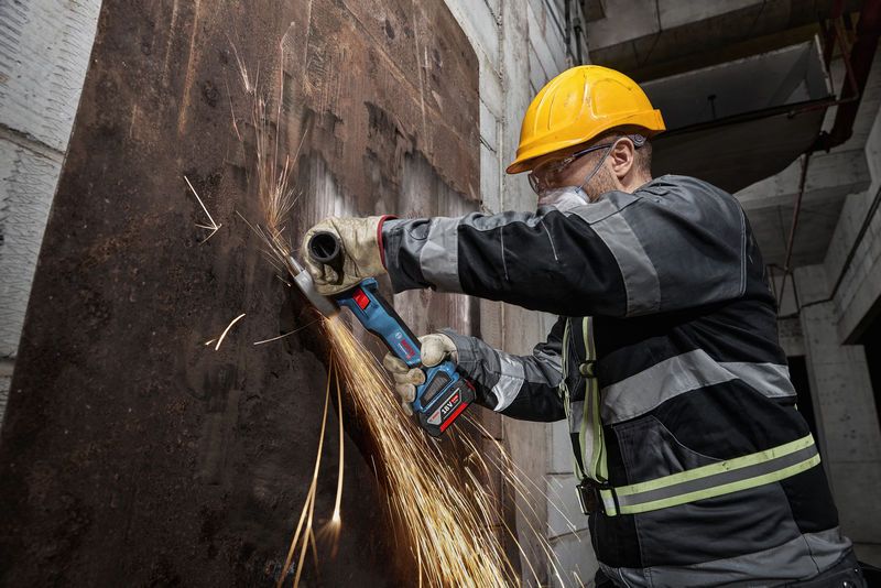 一名配戴安全設備的人正在用無線角磨機打磨生鏽的金屬板。.