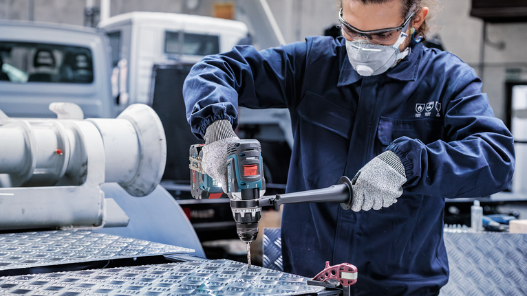 一名配戴安全設備的人員在工業環境中鑽入金屬板。