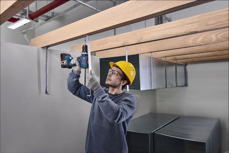 一名配戴安全設備的人員正在將鑽頭插入木樑下方的金屬棒。.