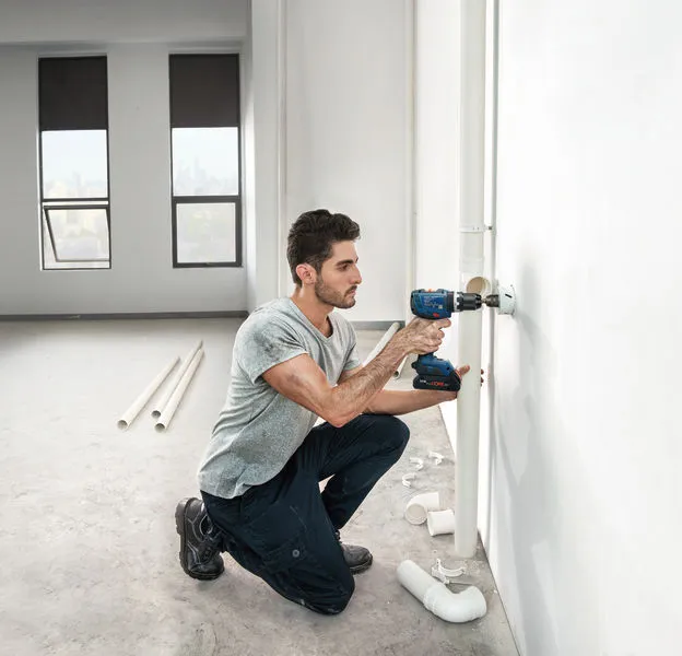 一個人在一間明亮的、未完工的房間裡鑽孔安裝管道。.