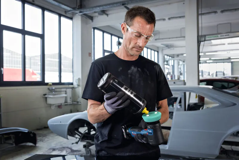 一名配戴安全設備的人員在汽車修理廠內對電動拋光機進行拋光。.