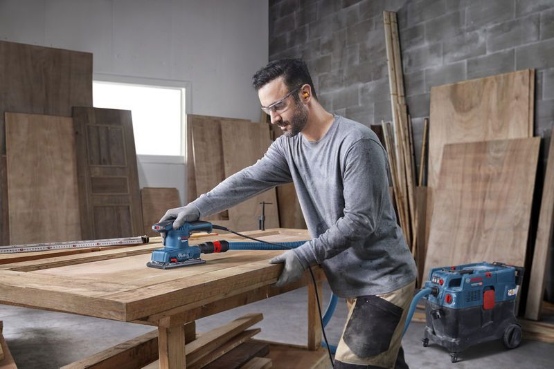 配戴安全設備的人員使用軌道砂光機打磨木質桌面。.