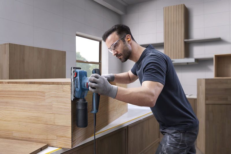 一名配戴安全設備的人員正在使用軌道砂光機打磨木製櫥櫃。.