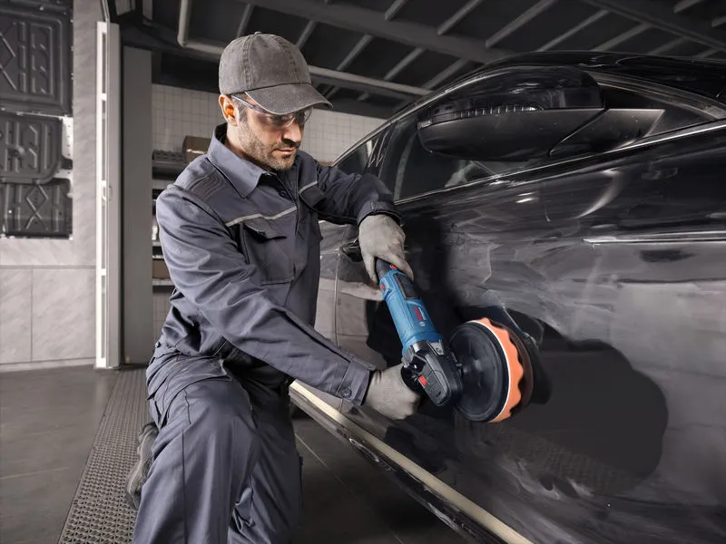 一名佩戴安全設備的人使用電動工具對一輛黑色汽車進行拋光。.