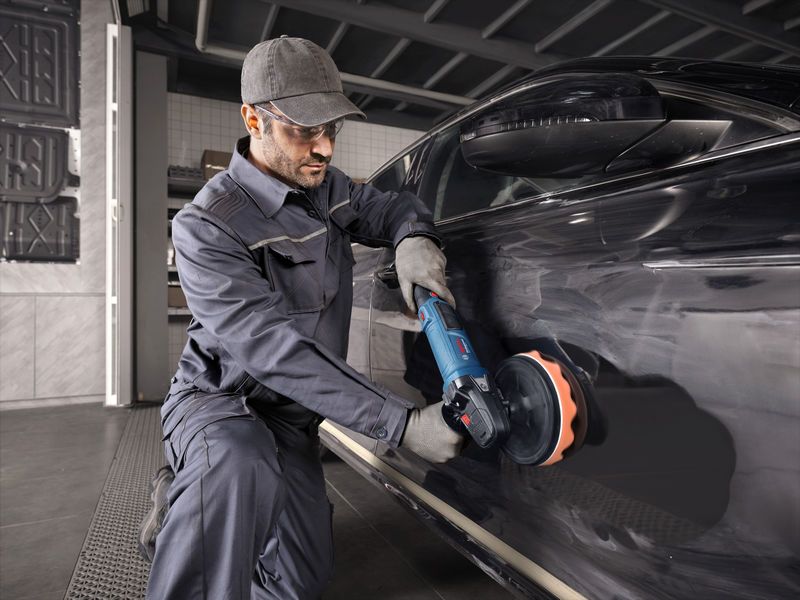 一名佩戴安全設備的人使用電動工具對一輛黑色汽車進行拋光。.