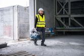 一名配戴安全設備的人員在一棟未完工的建築物內攜帶一把金屬切割鋸。