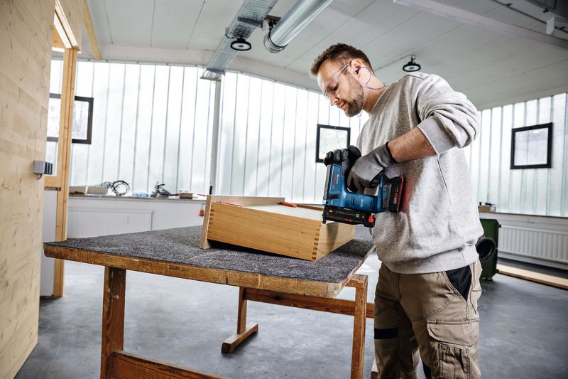 一名配戴安全設備的人員在車間使用無線釘槍敲擊木箱。.