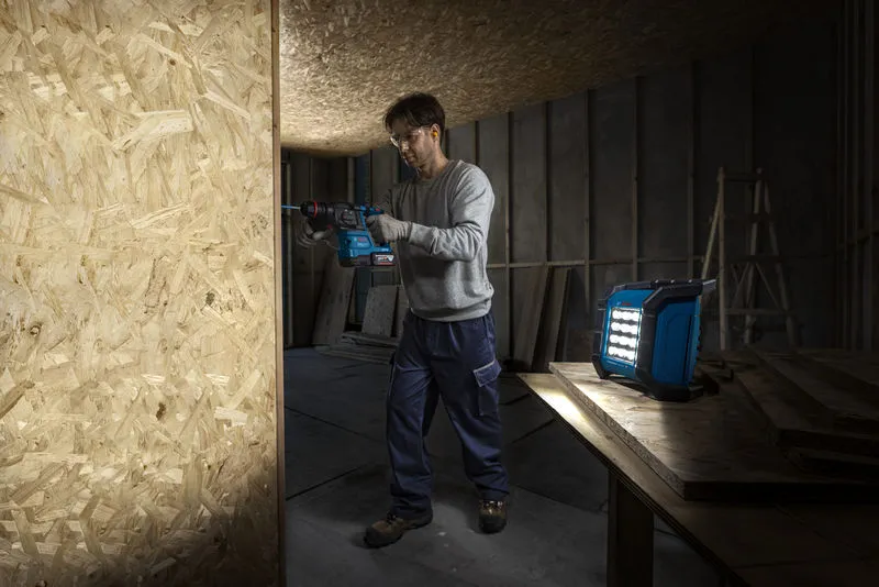 一名配戴安全設備的人員正在無線工地燈附近的木板上鑽孔。.