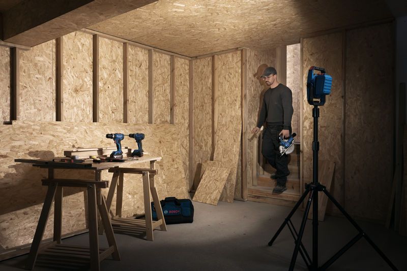 一名佩戴安全裝備的人走進由無線工地燈照亮的木工車間。.