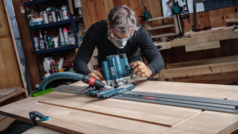 配戴安全設備的人員引導路由器沿著軌道塑造木板。.