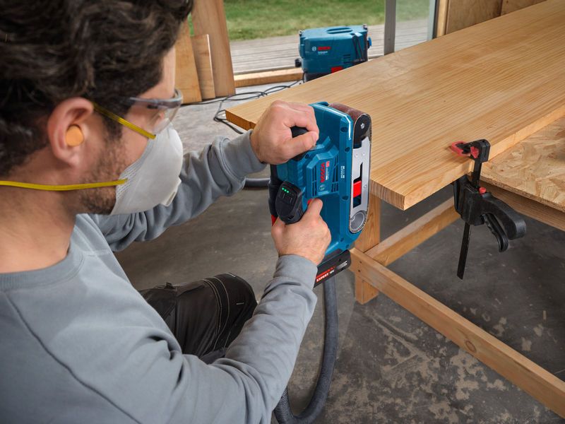 一名穿戴安全裝備的人員正在使用帶式砂光機打磨木板。