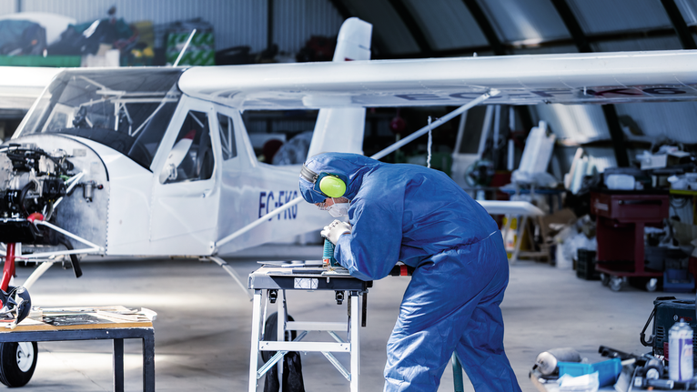 一名配戴安全設備的人員在小型飛機附近的工作台上鑽金屬零件。.