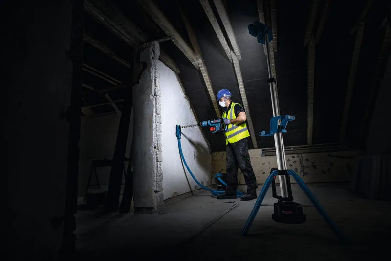 一名穿戴安全裝備的人員正在牆上鑽孔，一盞無線工地照明燈照亮了牆面。