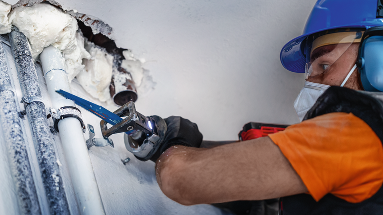 一名穿戴安全裝備的人員使用往復鋸切割靠近牆壁的管道。